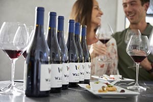 Bottles of WALT Pinot Noir on a table with a couple in the background with glasses of red wine in their hands
